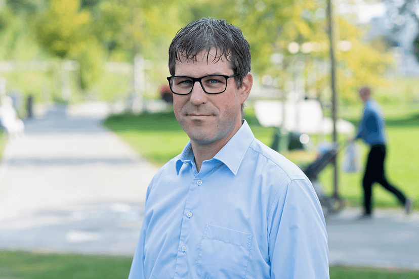 Michael Degiampietro, a person in glasses and a light blue shirt stands in a park, with blurred greenery and a person pushing a stroller in the background.