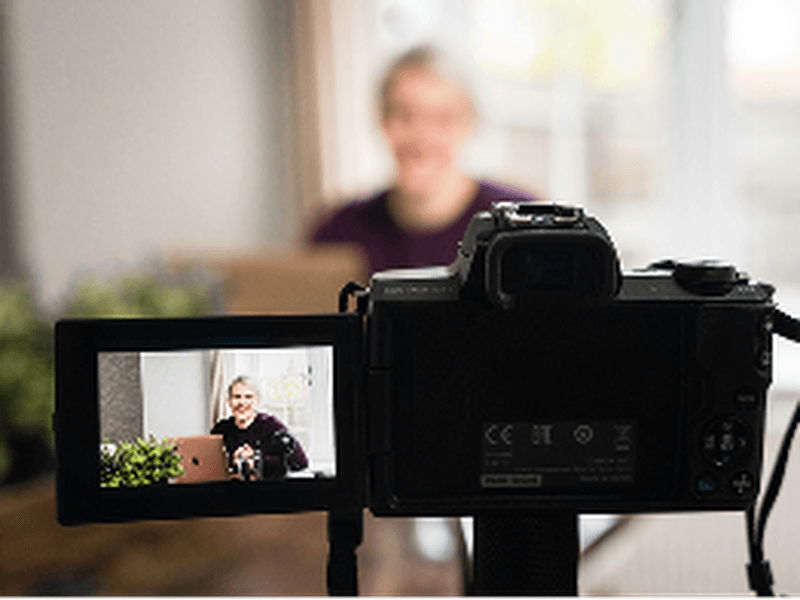 A digital camera on a tripod records a person sitting indoors, with the camera screen showing their image and a blurred background.
