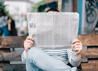 Person sitting on a bench and reading a newspaper, wearing a striped shirt and jeans. The background is slightly blurred with an urban setting.