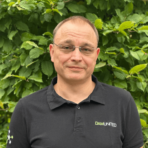 A person wearing glasses and a black shirt with "DAMUNITED" embroidered stands in front of green leafy bushes.